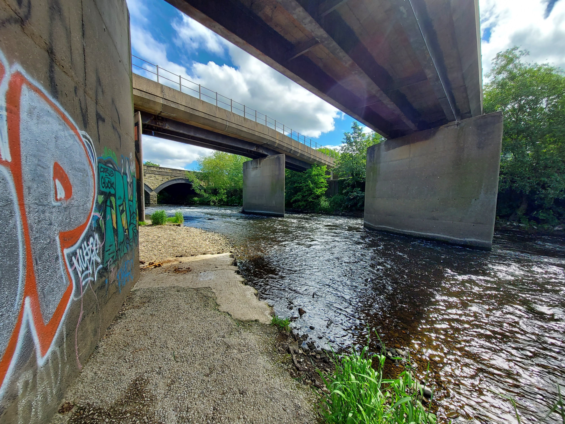 Heavy concrete bridge piers standing in a river beneath a modern road bridge, with an older stone arch bridge behind.