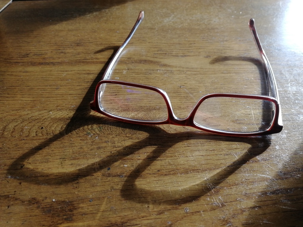 A pair of reading glasses resting on a wooden table in sharp afternoon light.