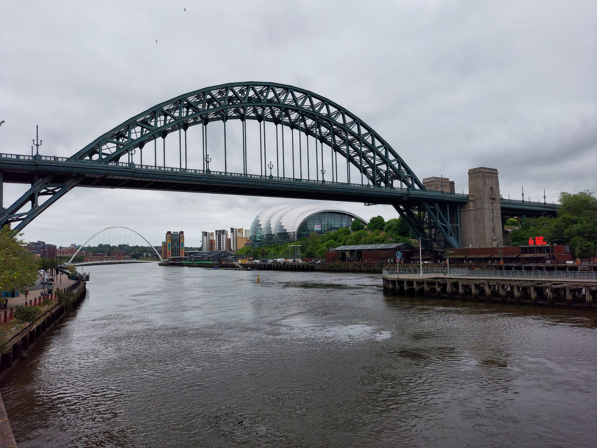 A large steel arch bridge spanning a river, with a second bridge and riverside buildings beyond.